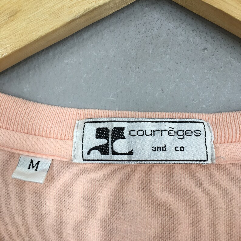 K&ouml;nnte beinhalten: Nahaufnahme eines pfirsichfarbenen Hemdes mit geripptem Ausschnitt. Das Etikett lautet "courr&egrave;ges and co" in einem rechteckigen wei&szlig;en Etikett mit schwarzem Logo. Ein kleineres Etikett mit dem Buchstaben "M" ist ebenfalls sichtbar.