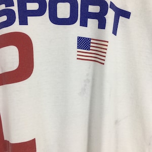 May include: White t-shirt with the word "SPORT" in blue letters and a red number "2" on the front. A small American flag is printed on the right side of the shirt.