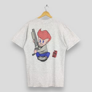 May include: Light grey t-shirt with a cartoon illustration of a boy with red hair holding a vacuum cleaner. The boy wears a grey shirt and blue trousers. A small red box is to the right of the boy. The t-shirt is hanging on a wooden hanger.
