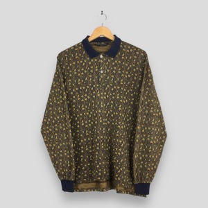 May include: A long-sleeved polo shirt with a dark navy blue collar and cuffs. The shirt has a pattern of small yellow and brown floral shapes on a dark gray background. The shirt is hanging on a wooden hanger.
