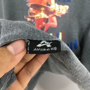 May include: Close-up of a gray t-shirt with a colorful graphic design. The shirt features a black tag with the Avirex logo. The shirt's fabric is visible, showing its texture and color.