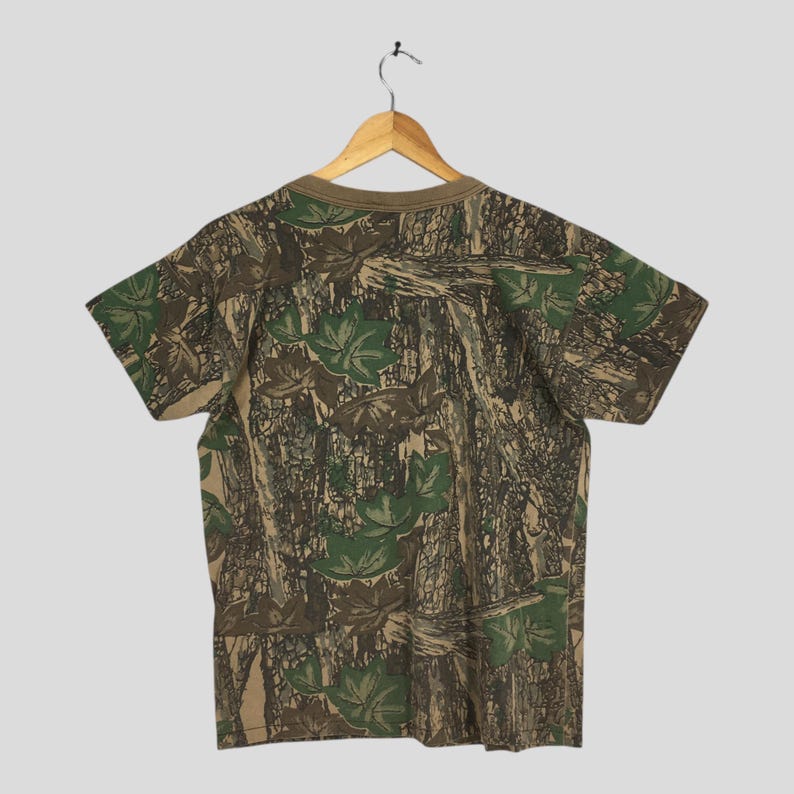 Puede incluir: Camiseta de manga corta con estampado de camuflaje. La camiseta presenta un cuello marr&oacute;n y un dise&ntilde;o de hojas verdes y corteza de &aacute;rbol. La camiseta est&aacute; colgada de una percha de madera sobre un fondo gris claro.