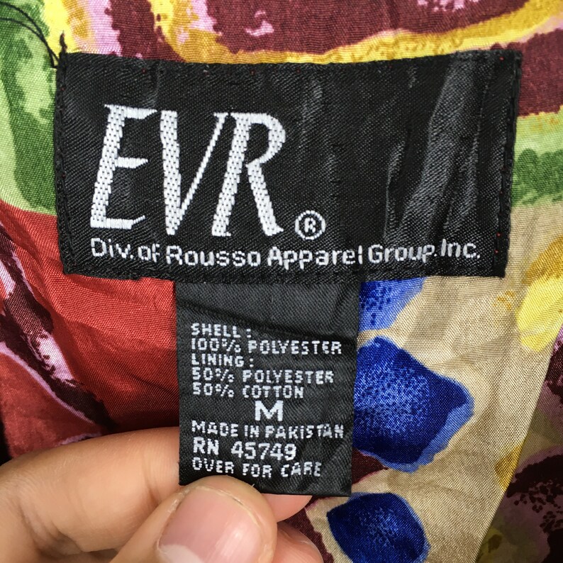 May include: A black and white clothing label with the text "EVR Div. of Rousso Apparel Group Inc." and care instructions for a garment. The label states that the shell is 100% polyester and the lining is 50% polyester and 50% cotton. The size is M. The garment was made in Pakistan.