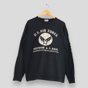 May include: Black long-sleeve shirt with the text "U.S. AIR FORCE" and a circular emblem. Below the emblem, the text reads "WESTOVER A. F. BASE MASSACHUSETTS". The shirt is hanging on a wooden hanger.