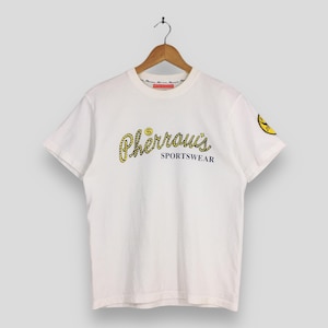 May include: White t-shirt with the text "Pherrous Sportswear" in yellow script. The shirt has a small yellow emblem on the sleeve. The shirt is hanging on a wooden hanger.