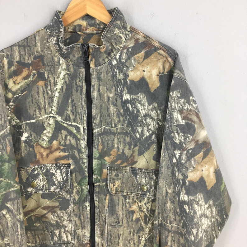 Op de afbeelding: Een camouflagejas met een ritssluiting en twee zakken. De jas is gemaakt van een dikke, duurzame stof en heeft een opstaande kraag.