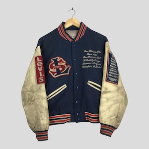 Puede incluir: Una chaqueta universitaria Levi's vintage azul marino con mangas de cuero color crema. La chaqueta presenta puños y cuello a rayas rojas y blancas, y varios parches con el logotipo y el texto de Levi's. La chaqueta está colgada de una percha de madera.