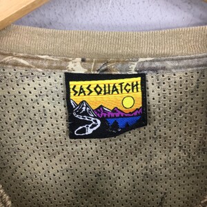 Pu&ograve; includere: Primo piano di una maglietta in rete beige con una toppa "Sasquatch". La toppa presenta una catena montuosa, un fiume, un sole e la scritta "Sasquatch". La maglietta ha un motivo mimetico sul colletto.