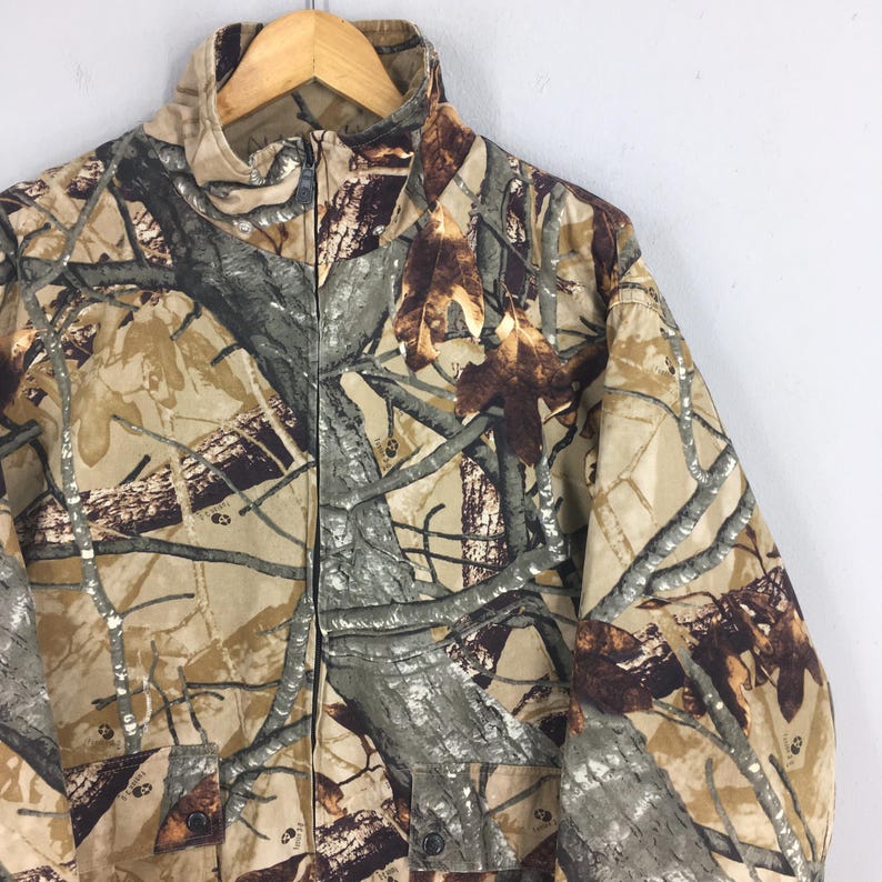 May include: A camouflage jacket with a full zipper closure and a high collar. The jacket features a pattern of brown, tan, and gray tree branches and leaves. It has two front pockets and is hanging on a wooden hanger.