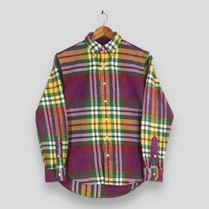 Puede incluir: Una camisa a cuadros con botones en una vibrante mezcla de colores, incluyendo morado, amarillo, verde y naranja. La camisa de manga larga presenta un cuello y puños clásicos, con un pequeño logo en el pecho. La camisa está colgada en una percha de madera.