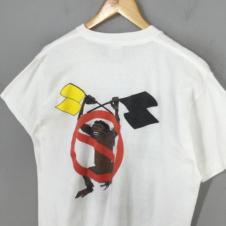 May include: White t-shirt featuring a graphic of a monkey holding yellow and black flags within a red prohibition sign. The monkey is brown, and the flags are yellow and black. The shirt is hanging on a wooden hanger.