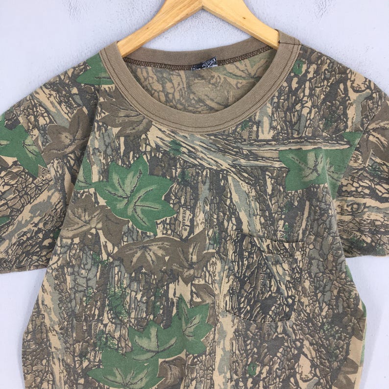 Puede incluir: Camiseta de camuflaje de manga corta con bolsillo en el pecho. La camiseta presenta un cuello marr&oacute;n y un estampado de hojas verdes y camuflaje marr&oacute;n y beige.