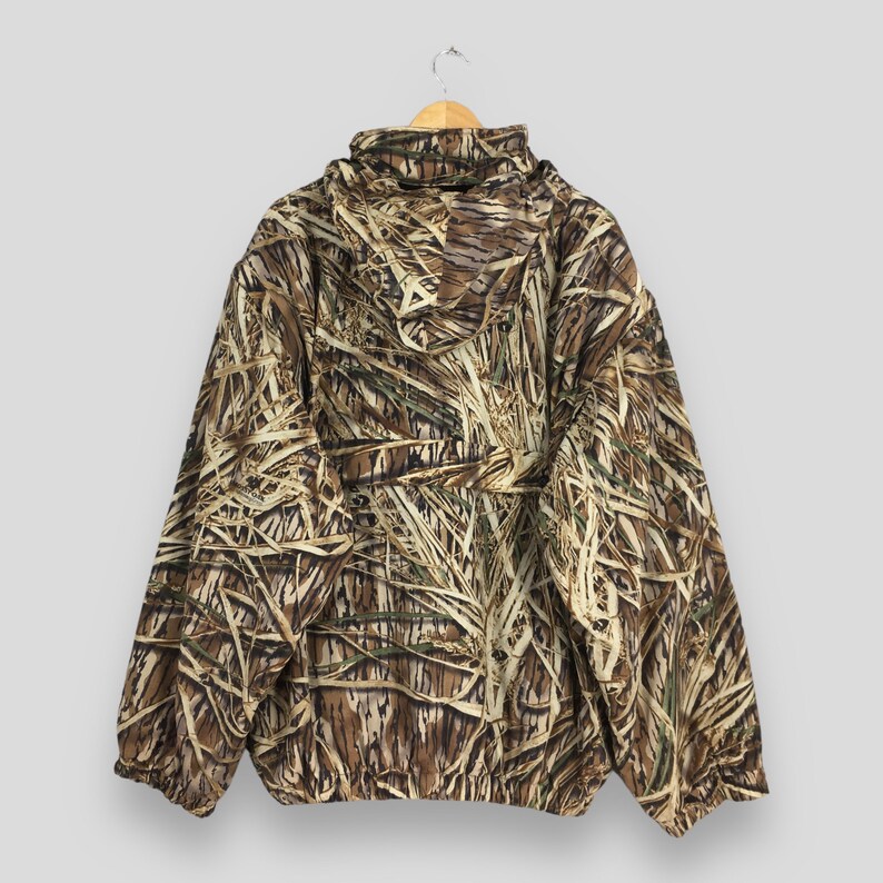 Puede incluir: Chaqueta de camuflaje con capucha, colgada de una percha de madera. La chaqueta presenta un patr&oacute;n de color marr&oacute;n, beige y verde que se asemeja a juncos y follaje. La chaqueta tiene cremallera y mangas largas.
