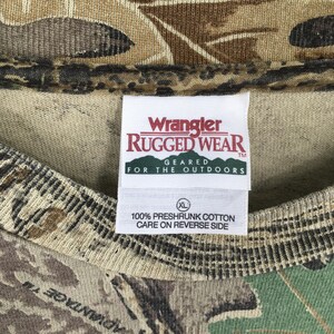以下が含まれることがあります： 迷彩柄のTシャツのクローズアップ。タグには赤と白で「Wrangler Rugged Wear」と書かれており、その下に「Geared for the Outdoors」と書かれています。タグには、シャツがXLサイズで、100％防縮加工の綿でできており、裏面に洗濯表示があることも記載されています。