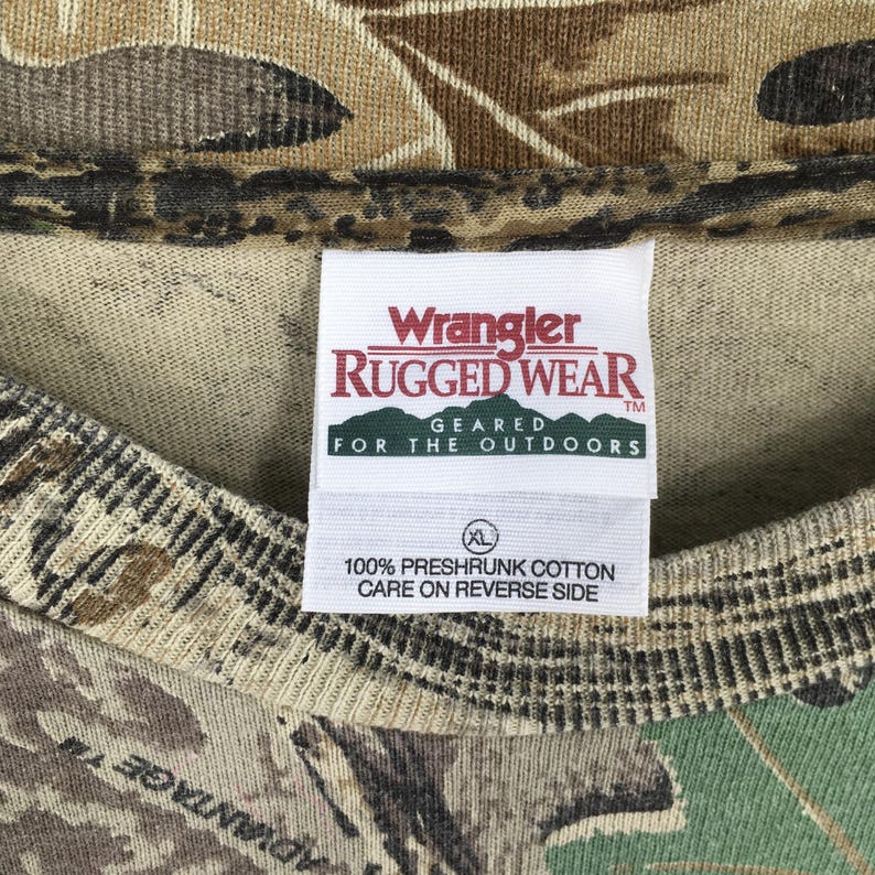 以下が含まれることがあります： 迷彩柄のTシャツのクローズアップ。タグには赤と白で「Wrangler Rugged Wear」と書かれており、その下に「Geared for the Outdoors」と書かれています。タグには、シャツがXLサイズで、100％防縮加工の綿でできており、裏面に洗濯表示があることも記載されています。