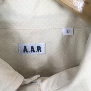 Pode incluir: Uma camisa branca de bot&otilde;es com um pequeno padr&atilde;o de bolinhas brancas. A camisa tem uma etiqueta que diz "A.A.R." e uma etiqueta de tamanho que diz "L".