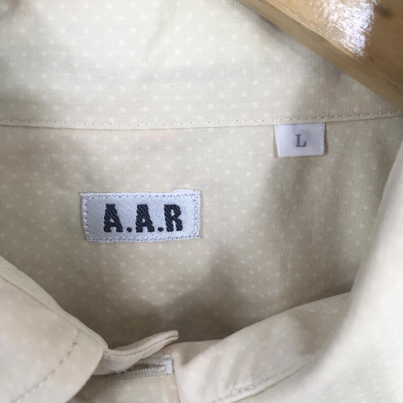 Pode incluir: Uma camisa branca de bot&otilde;es com um pequeno padr&atilde;o de bolinhas brancas. A camisa tem uma etiqueta que diz "A.A.R." e uma etiqueta de tamanho que diz "L".