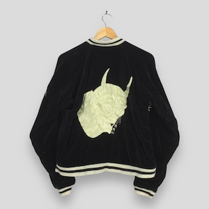 May include: Black bomber jacket featuring a cream-coloured embroidered demon face on the back. The jacket has cream-coloured stripes on the collar, cuffs, and hem. The jacket is hanging on a wooden hanger.