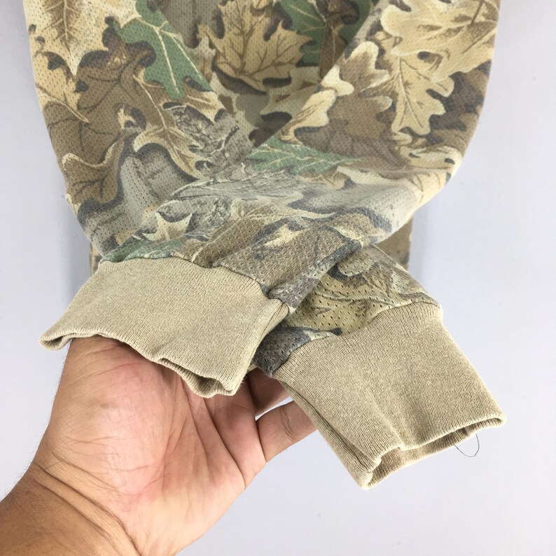 Pu&ograve; includere: Primo piano di una camicia a maniche lunghe mimetica. La camicia presenta un motivo di foglie marroni, verdi e beige. I polsini sono di colore beige pieno. Il tessuto sembra essere un materiale a rete traspirante.