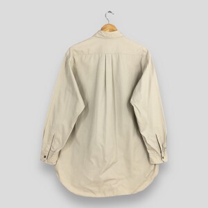 Peut inclure: Chemise beige &agrave; manches longues, suspendue &agrave; un cintre en bois. Elle pr&eacute;sente un col classique, des poignets boutonn&eacute;s et un ourlet arrondi. Le dos de la chemise r&eacute;v&egrave;le un pli vertical. Tissu de couleur claire.