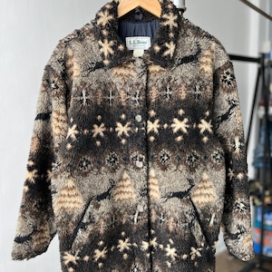 May include: An L.L.Bean long-sleeve jacket made of a textured, fuzzy fleece material. The jacket features a dark brown, tan, and cream-colored pattern with repeating snowflake and tree motifs, a collared neckline, and a front button closure.