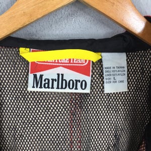 May include: A close-up of a black and red jacket with a yellow tag. The jacket features a Marlboro logo on a red and white tag. The tag includes text that reads "MADE IN TAIWAN" and "SIZE: L". The jacket is made of nylon.