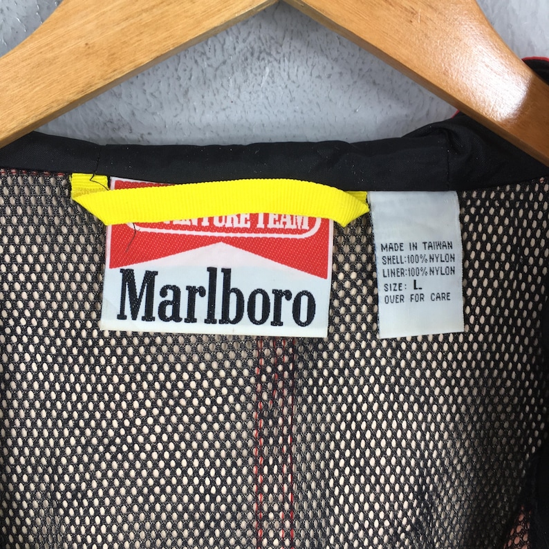 May include: A close-up of a black and red jacket with a yellow tag. The jacket features a Marlboro logo on a red and white tag. The tag includes text that reads "MADE IN TAIWAN" and "SIZE: L". The jacket is made of nylon.