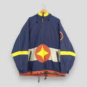 May include: A blue and yellow anorak jacket with a starburst design on the front. The jacket has a hood, a half-zip closure, and a patch on the sleeve. The jacket is hanging on a wooden hanger.