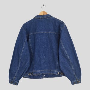 May include: A vintage denim jacket in a medium wash blue, hanging on a wooden hanger. The jacket features a classic collar, button closures, and pockets. The back view showcases the jacket's tailored design and seams.