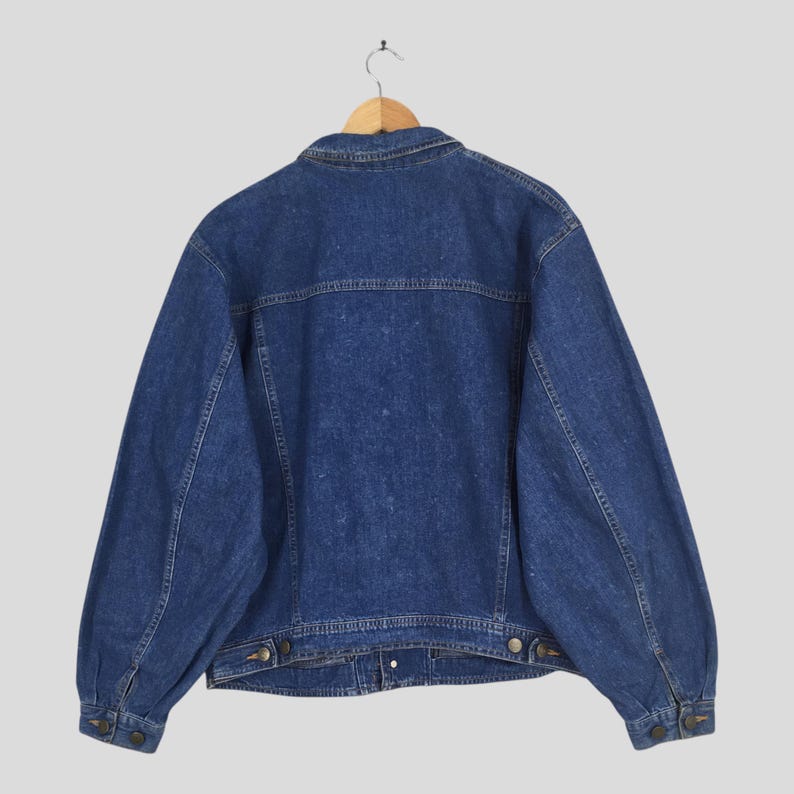 May include: A vintage denim jacket in a medium wash blue, hanging on a wooden hanger. The jacket features a classic collar, button closures, and pockets. The back view showcases the jacket's tailored design and seams.