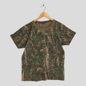 Puede incluir: Una camiseta de manga corta con estampado de camuflaje. La camiseta tiene cuello redondo y un cuello marr&oacute;n. El dise&ntilde;o de camuflaje incorpora tonos verdes, marrones y beige, con patrones de hojas y corteza de &aacute;rbol. Cuelga de una percha de madera.