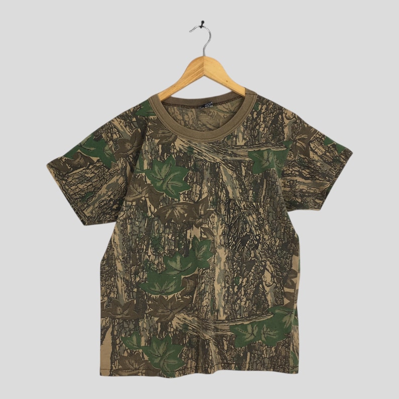 Puede incluir: Una camiseta de manga corta con estampado de camuflaje. La camiseta tiene cuello redondo y un cuello marr&oacute;n. El dise&ntilde;o de camuflaje incorpora tonos verdes, marrones y beige, con patrones de hojas y corteza de &aacute;rbol. Cuelga de una percha de madera.