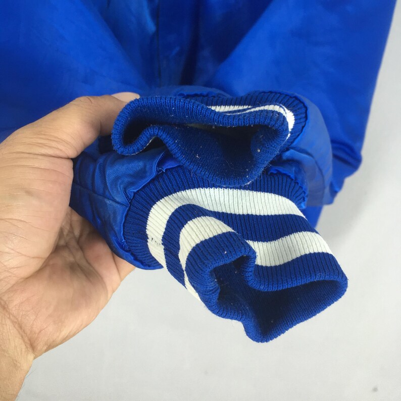 May include: A bright blue satin jacket sleeve featuring a ribbed knit cuff. The cuff is designed with a solid blue base and two horizontal white stripes running around the circumference, providing a distinct contrast to the smooth blue fabric.