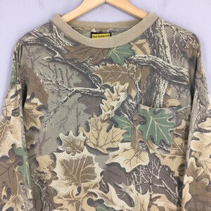 Pu&ograve; includere: Maglietta mimetica a maniche lunghe con tasca. La maglietta presenta un motivo di foglie e rami nei toni del marrone, verde e beige. Il colletto e i polsini sono di colore beige tinta unita. L'etichetta recita "SASQUATCH".