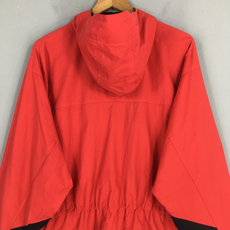 May include: A vibrant red hooded jacket with a drawstring waist and black detailing. The jacket is made of a lightweight material and has a full zip. The hood is attached and the jacket is hanging on a wooden hanger.