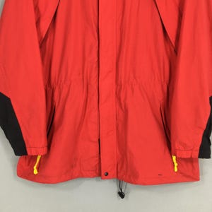 May include: A vibrant red jacket with black panels on the sleeves. The jacket features a drawstring waist and yellow detailing on the pockets and hem. The material appears to be lightweight.