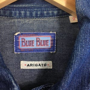 May include: Close-up of a blue denim shirt with a blue and white label that reads "Blue Blue" and a white label that reads "ARIGATO".
