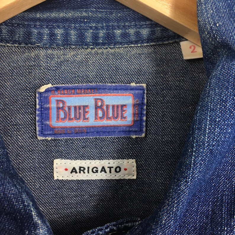 May include: Close-up of a blue denim shirt with a blue and white label that reads "Blue Blue" and a white label that reads "ARIGATO".