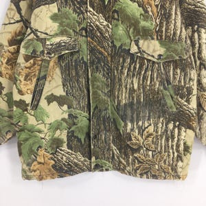 May include: Camouflage jacket featuring a pattern of green leaves and brown tree bark. The jacket has two front pockets with button closures and a front zipper. The fabric is a blend of green, brown, and tan colours.