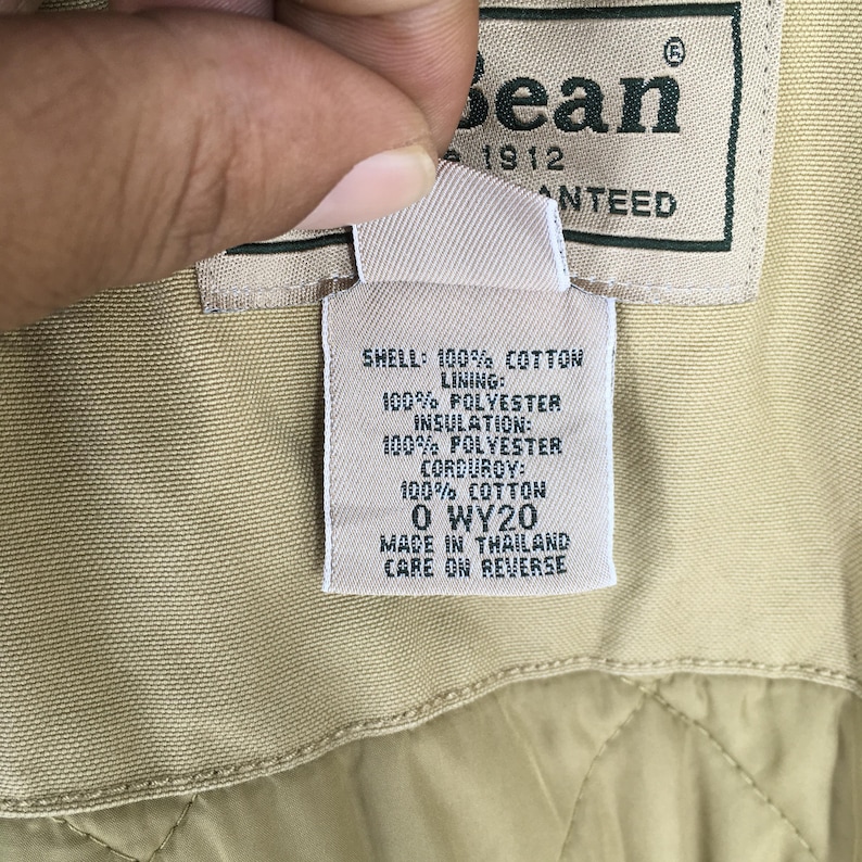 May include: Close-up of a tan jacket tag with the brand name "Bean" and the year "1912". The tag details the jacket's materials: shell 100% cotton, lining 100% polyester, insulation 100% polyester, and corduroy 100% cotton. The tag also includes the code "O WY20".