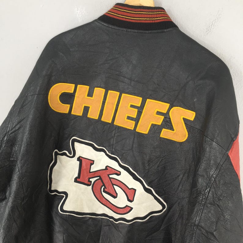 May include: Black leather jacket with the word "CHIEFS" in yellow block letters. Below the word is a white and red logo. The jacket has a red accent on the side and a striped collar.