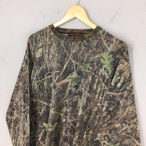 May include: Long-sleeved camouflage shirt with a crew neck. The shirt features a brown, green, and tan pattern designed to blend into a wooded environment. The shirt is hanging on a wooden hanger.