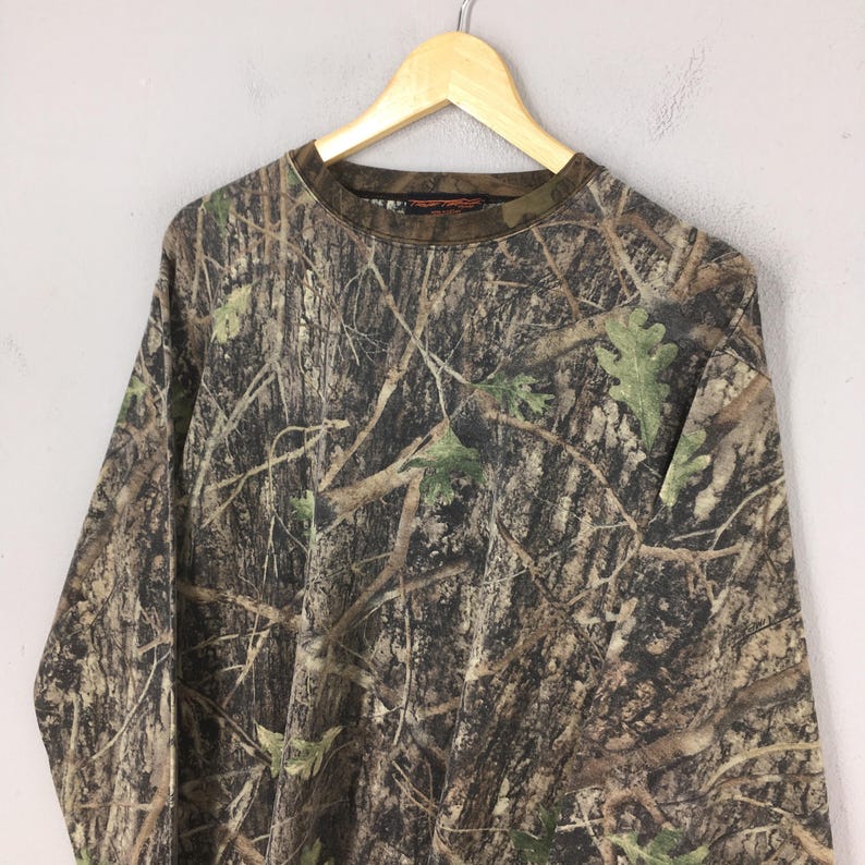 May include: Long-sleeved camouflage shirt with a crew neck. The shirt features a brown, green, and tan pattern designed to blend into a wooded environment. The shirt is hanging on a wooden hanger.