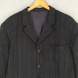 May include: A dark charcoal gray blazer featuring a subtle vertical striped pattern, a notched lapel collar, and two black buttons on the front closure. The garment includes a single chest pocket and a smooth dark interior lining.