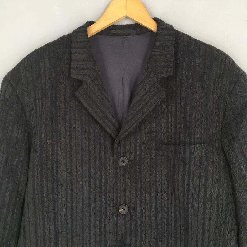 May include: A dark charcoal gray blazer featuring a subtle vertical striped pattern, a notched lapel collar, and two black buttons on the front closure. The garment includes a single chest pocket and a smooth dark interior lining.