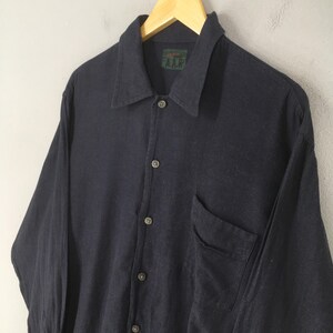 Puede incluir: Una camisa abotonada de color azul marino oscuro con mangas largas y un bolsillo en el pecho. La camisa est&aacute; colgada en una percha de madera. El cuello tiene una peque&ntilde;a etiqueta verde con texto blanco. La camisa es de tela texturizada.