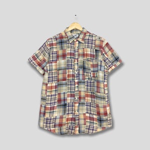 Pode incluir: Camisa de manga curta com patchwork em vários padrões xadrez. A camisa tem uma frente abotoada, uma gola e um bolso no peito. A paleta de cores inclui vermelho, azul, amarelo e creme.