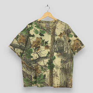 May include: A short-sleeved t-shirt with a camouflage pattern featuring shades of green, brown, and beige, resembling a forest scene. The shirt is hanging on a wooden hanger.