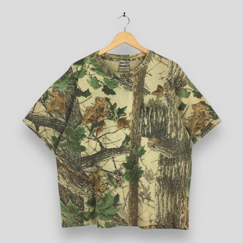 May include: A short-sleeved t-shirt with a camouflage pattern featuring shades of green, brown, and beige, resembling a forest scene. The shirt is hanging on a wooden hanger.