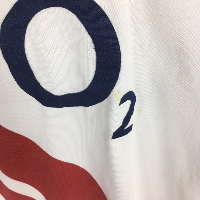 Peut inclure: Gros plan sur un tissu blanc avec un grand "O" bleu marine et un "2" bleu marine imprim&eacute;s. Un motif rouge et blanc est visible en bas. Le tissu a une l&eacute;g&egrave;re texture.